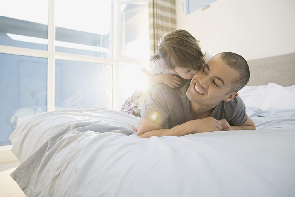Ein Vater liegt im Bett mit seiner Tochter auf dem Rücken und sie gibt ihm einen Kuss auf die Backe.