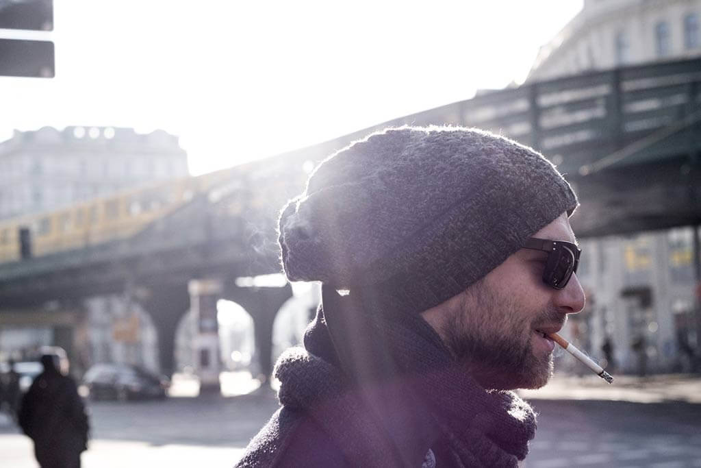 Ein Mann mit Bart trägt eine Wollmütze und eine Sonnenbrille und raucht eine Zigarette.