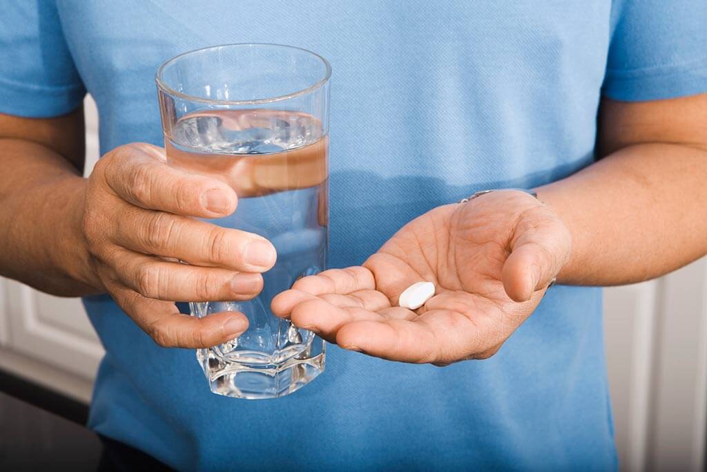 Ein Mann hält in seiner linken Hand ein Glas Wasser und in der anderen Hand eine Tablette.