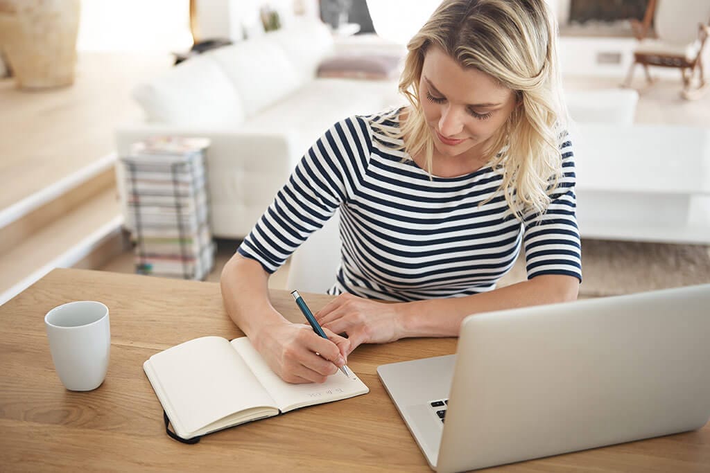 Junge Frau sitzt am Tisch mit ihrem Laptop und notiert sich etwas in ein Notizbuch.