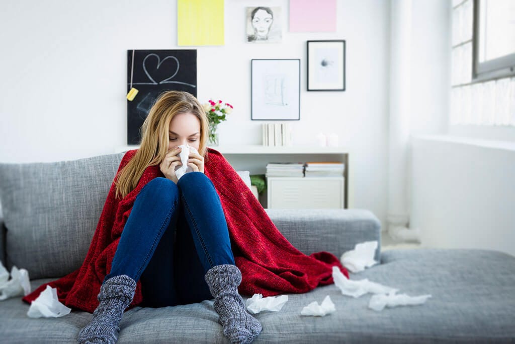 Junge Frau sitzt verschnupft mit vielen Taschentüchern auf einem Sofa.