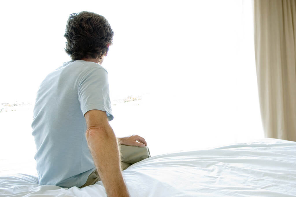 Ein Mann sitzt auf einem Bett und schaut aus einem Fenster.