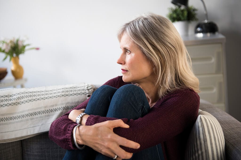 Eine Frau sitzt auf dem Sofa, schaut sehr nachdenklich und umklammert ihre Beine.
