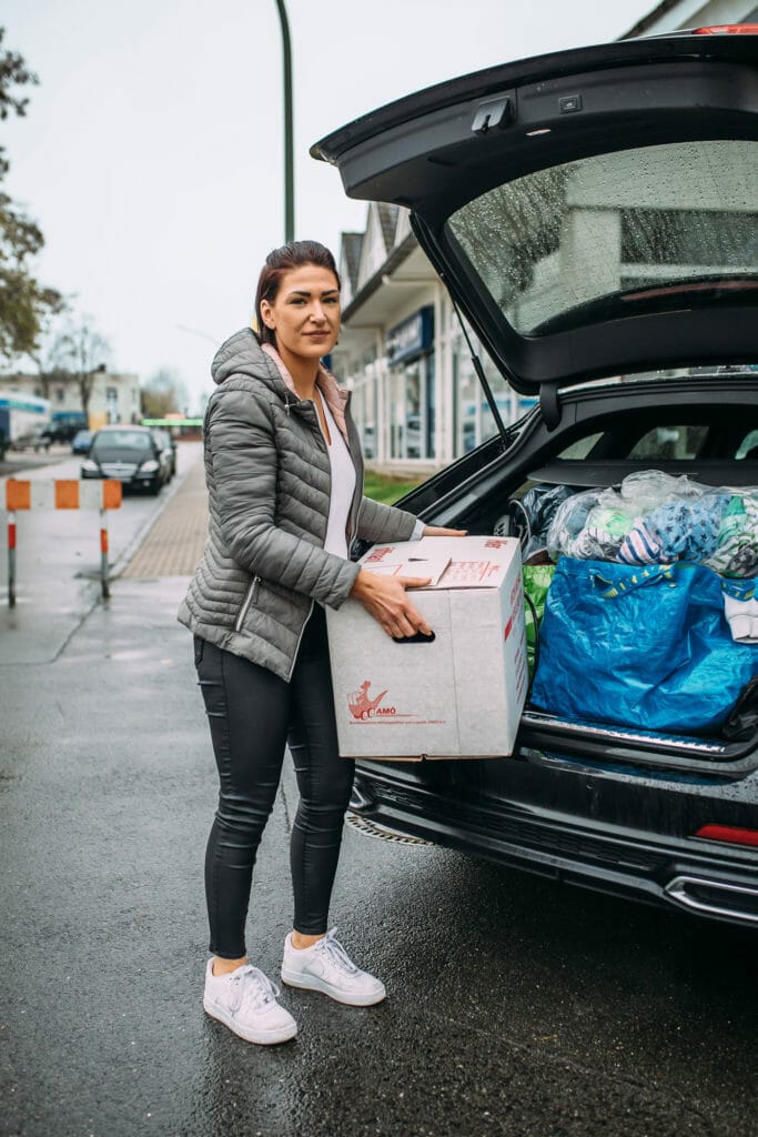 Germaine steht mit einem Umzugskarton vor Ihrem gepackten Auto.