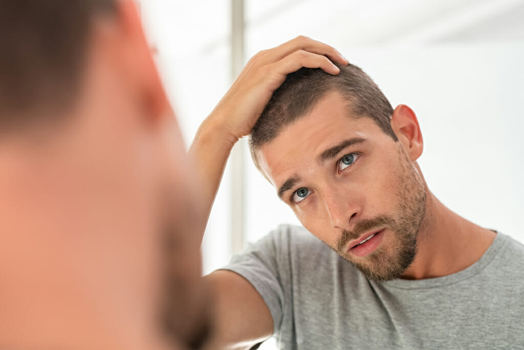 Ein junger Mann schaut sich im Spiegel seine Kopfhaut an.