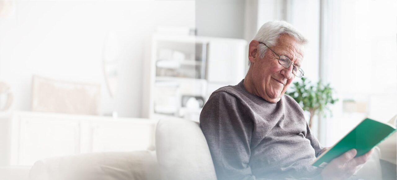 Ein älterer Mann mit weißen Haaren und einer Brille liest ein Buch auf dem Sofa