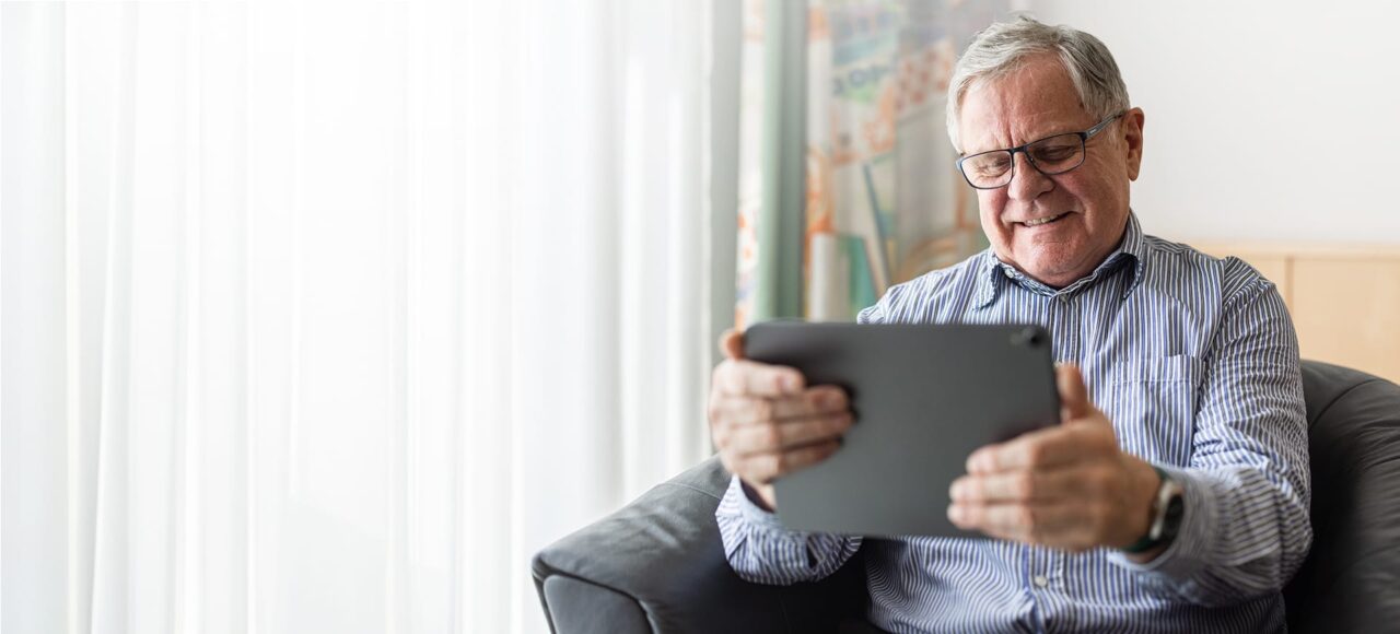 Älterer Mann mit Hemd und Brille schaut auf sein Tablet und lächelt