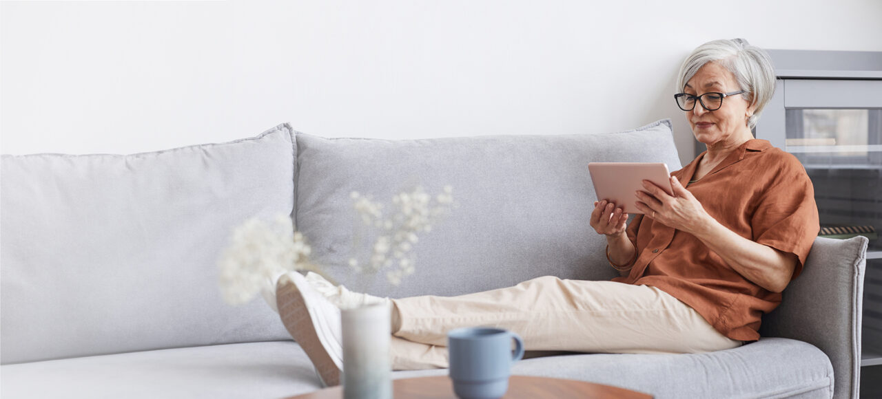 Frau mit grauen Haaren und Brille sitzt auf dem Sofa und schaut etwas auf dem Tablet an