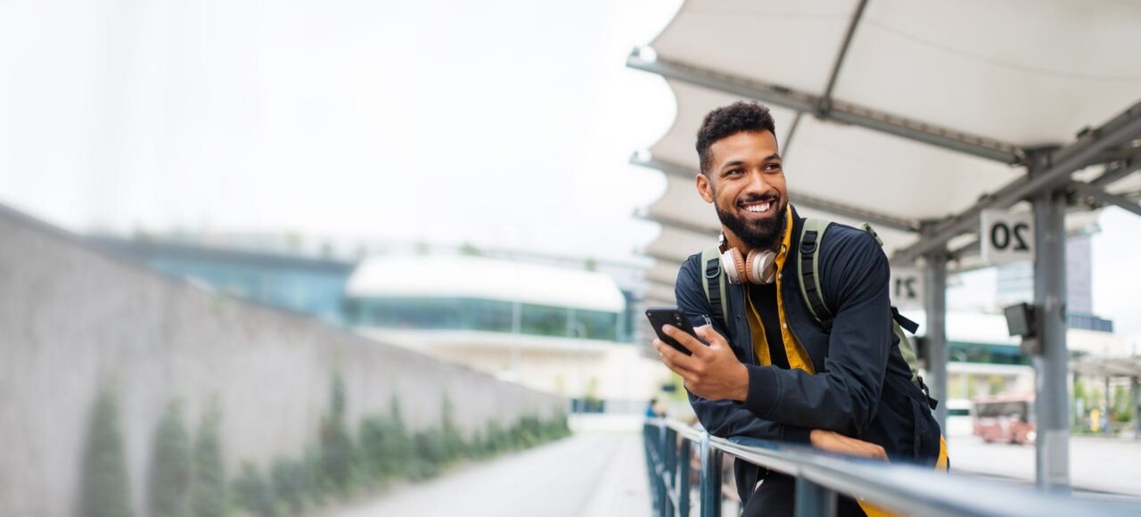 Junger Mann an einem Geländer angelehnt, hält lachend, sein Handy in der Hand.