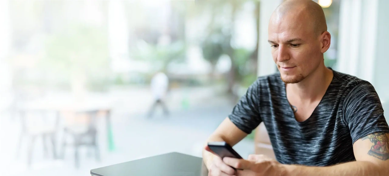 Mann mit Glatze sitzt am Schreibtisch und recherchiert am Handy