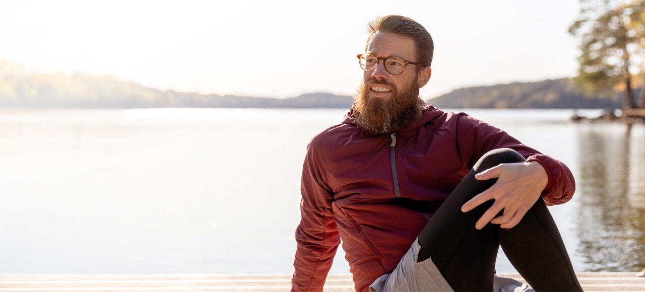 Mann mit Bart und in Sportkleidung sitzt am See und dehnt sich