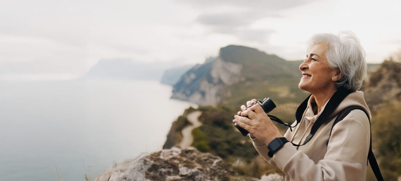 Ältere Frau mit grauen kurzen Haaren steht an einer Klippe vor dem Meer und schaut durchs Fernglas