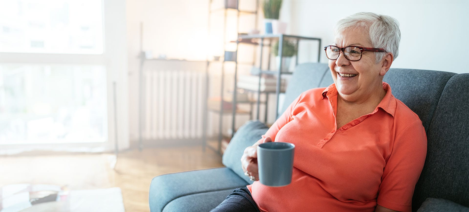 Ältere Frau mit grauen Haaren und Brille trinkt einen Kaffee auf dem Sofa