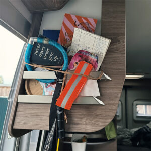 Ein kleines Regal in einem Camper ist mit Notizzetteln, einem Schlüsselband und weiteren Reiseutensilien gefüllt, die von einem Gummiband gesichert werden