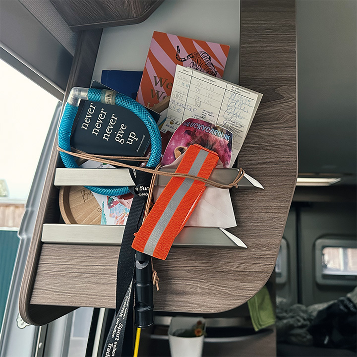 Ein kleines Regal in einem Camper ist mit Notizzetteln, einem Schlüsselband und weiteren Reiseutensilien gefüllt, die von einem Gummiband gesichert werden