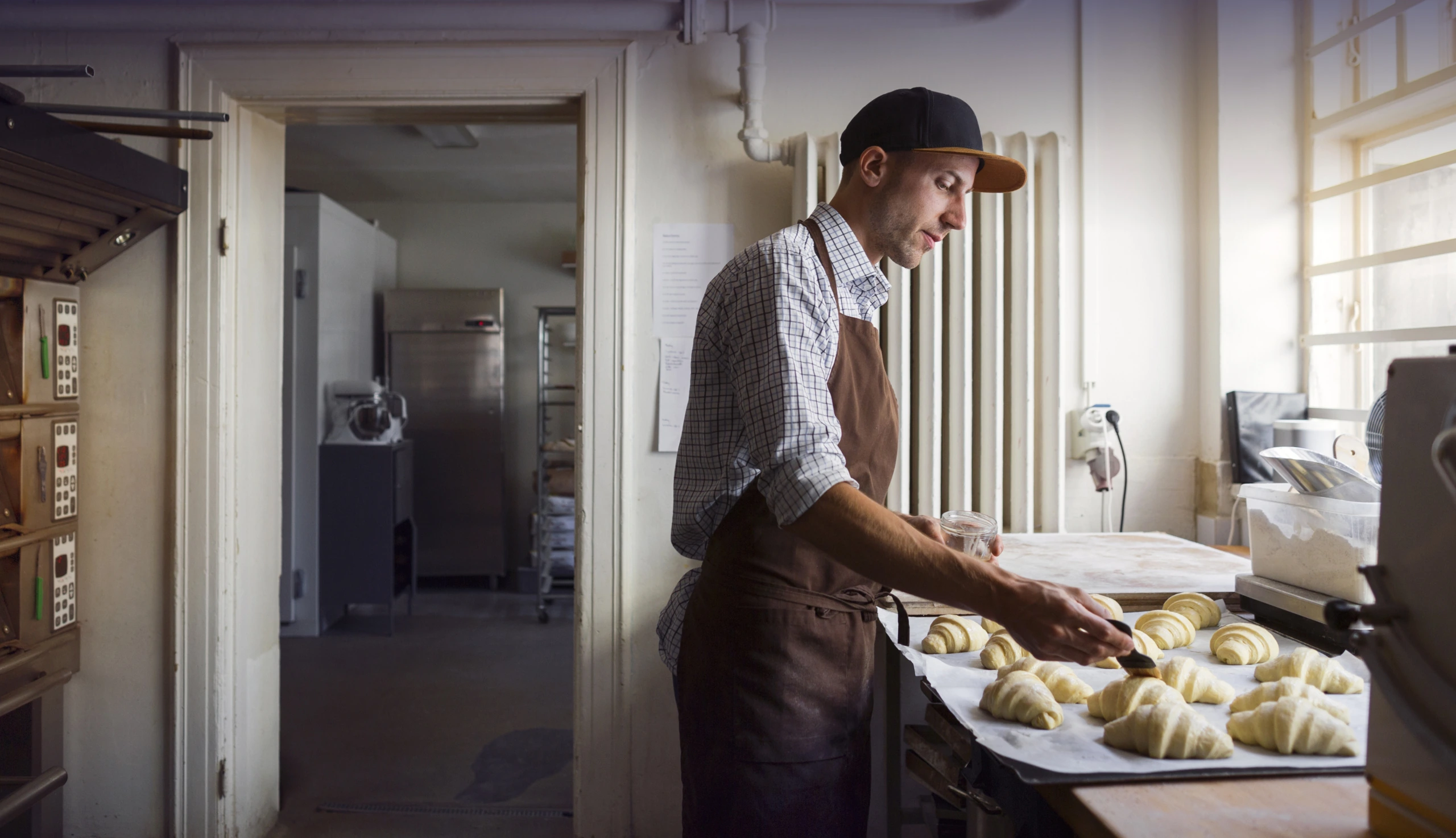 Junger Mann mit Mütze steht in der Küche, hat eine Schürze an und kocht.
