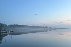 Ruhiger See in der Morgendämmerung mit leichtem Nebel, stiller Wasseroberfläche, Uferbäumen und einigen kleinen Booten am linken Bildrand.
