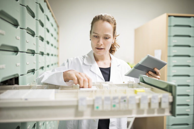 Apothekerin im weißen Kittel steht vor einem Medikamentenschrank und greift nach einer Packung.