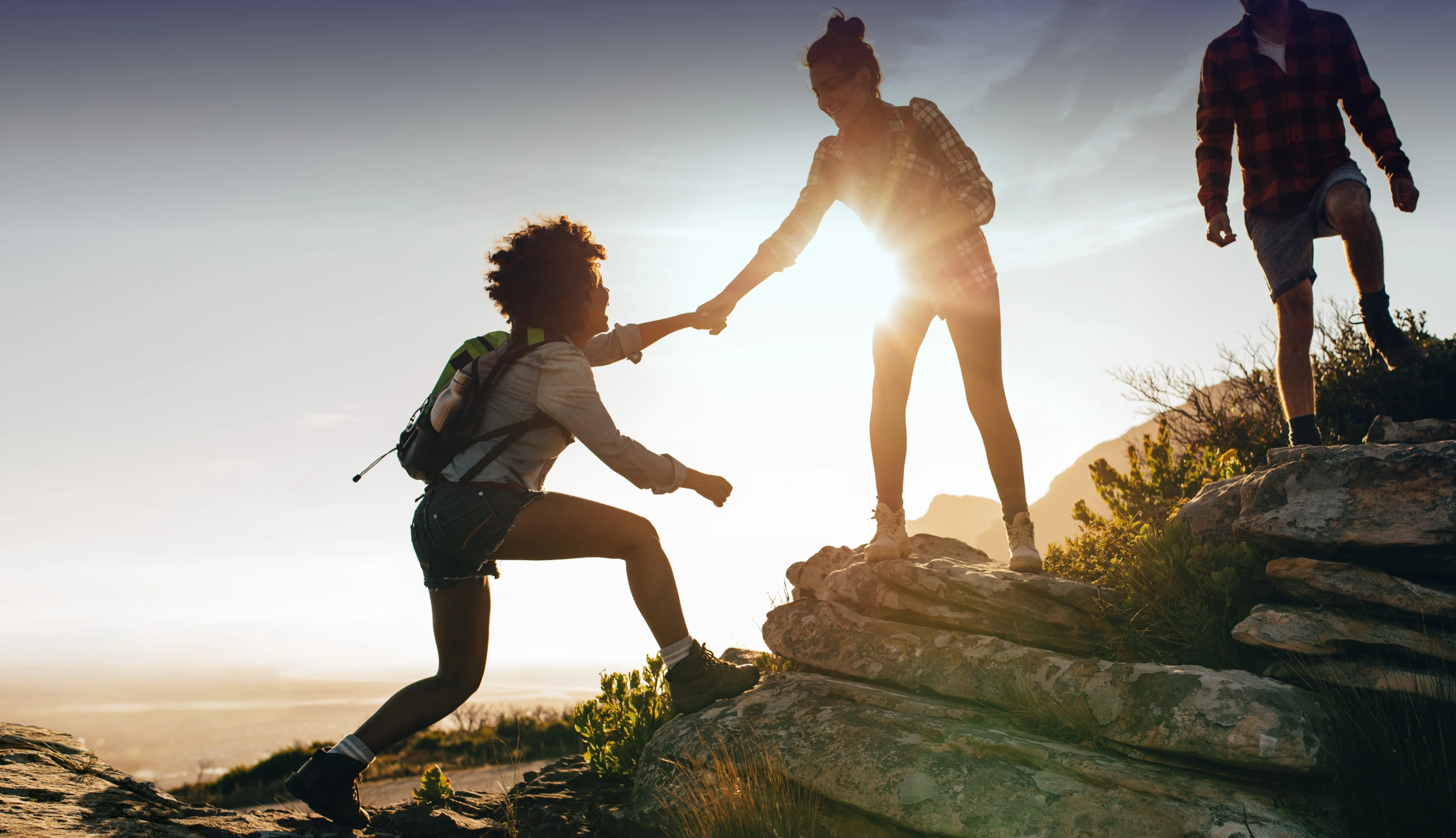 Zwei Frauen und ein Mann bei einer Wanderung. Die Frauen helfen sich gegenseitig beim Aufstieg.