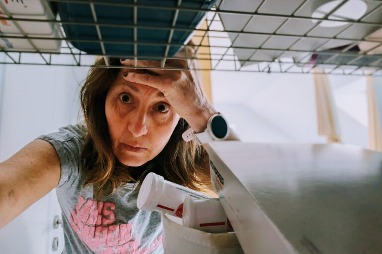 Frau mit Migräne greift erschöpft in einen Medikamentenschrank und hält sich die Stirn.