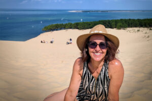 Tanja sitzt mit einem Sonnenhut und einer Sonnenbrille am Strand.